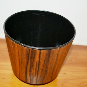 May include: A decorative planter with a wood grain finish. The exterior has a brown and black pattern, while the interior is black. The planter has a slightly tapered shape and is empty.