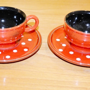 May include: Two red teacups with white polka dots and black interiors, each sitting on a matching saucer.