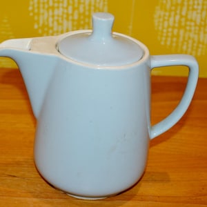 May include: A light blue ceramic teapot with a white lid and a handle. The teapot is on a wooden surface.
