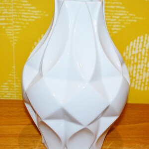 May include: A white ceramic vase with a geometric design. The vase has a wide base and a narrow neck.