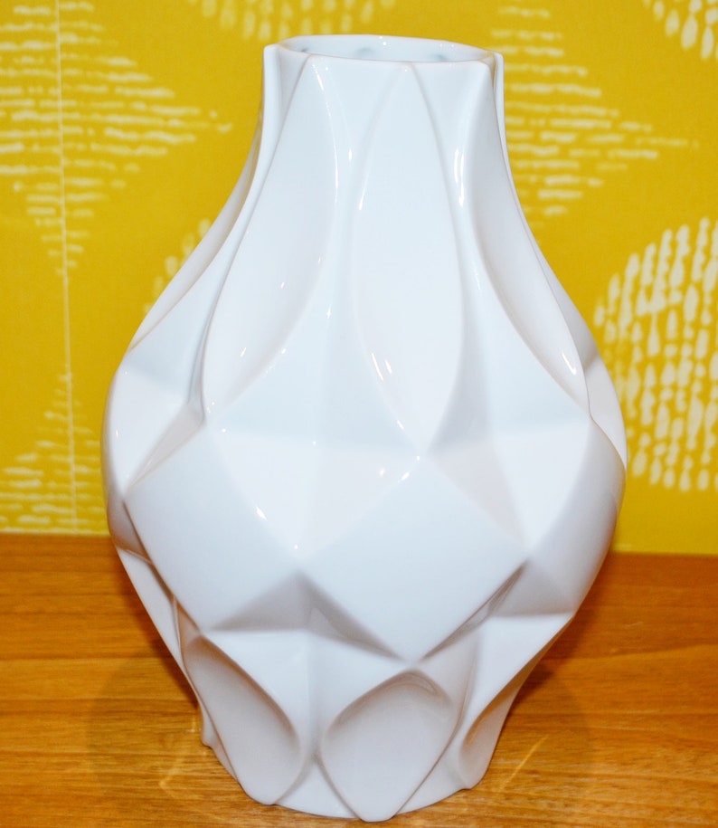 May include: A white ceramic vase with a geometric design. The vase has a wide base and a narrow neck.