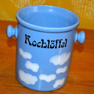 Puede incluir: Una olla de cerámica azul claro con dos asas laterales y la palabra "Kochlöffel" en escritura negra. Formas de nubes blancas están pintadas alrededor del exterior de la olla. La olla es probablemente para uso en la cocina.