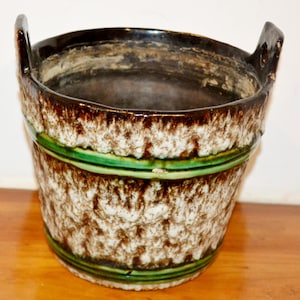 Può includere: Un vaso in ceramica marrone e bianco con strisce verdi e due manici. Il vaso ha una superficie testurizzata e un aspetto rustico.