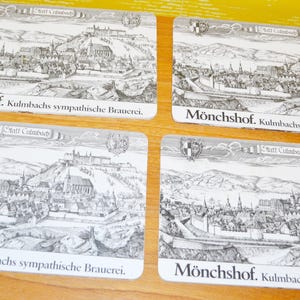 Pode incluir: Quatro bases de copos retangulares com uma ilustração a preto e branco de uma paisagem urbana, com uma montanha e edifícios. As bases de copos têm o texto "Mönchshof. Kulmbachs sympathische Brauerei."