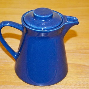 Puede incluir: Una jarra de cerámica azul cobalto con tapa y asa. La jarra tiene un asa curva y un pico vertedor. La tapa tiene un pequeño pomo en la parte superior. La jarra está sobre una superficie de madera.