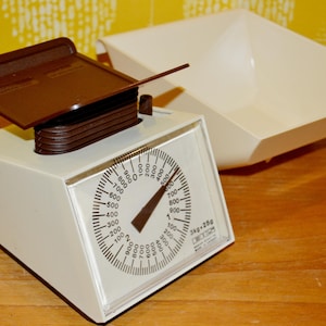 May include: A retro kitchen scale with a white body, brown weighing platform, and a dial face. The scale has a capacity of 3 kg or 25g. A white, square bowl is next to the scale. The scale is on a wooden surface.