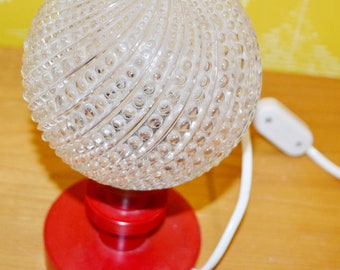 May include: A vintage table lamp with a clear glass globe shade featuring a textured, bubbled design. The shade is mounted on a red base with a white cord and switch.