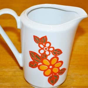 May include: White ceramic milk jug with a floral design. The flowers are orange, yellow, and white with brown leaves.