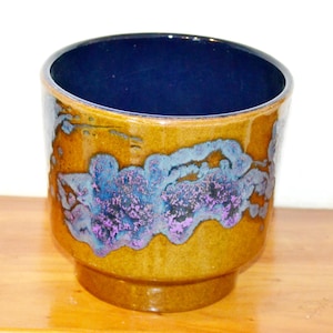May include: A ceramic planter with a dark blue interior and a textured exterior. The exterior features a blend of brown, blue, and purple hues. The planter has a rounded shape with a pedestal base.