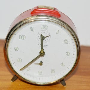 Könnte beinhalten: Ein runder roter analoger Wecker mit weißem Zifferblatt, goldfarbenem Metallrand und zwei kleinen Standfüßen. Die Uhr zeigt schwarze Ziffern, ein zentrales Wecker-Hilfszifferblatt und den Aufdruck Junghans Trivox mit drei Metallzeigern.