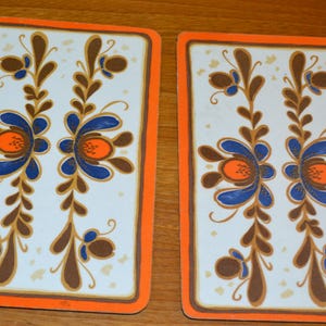 Pode incluir: Dois  placemats vintage com fundo branco e borda laranja. Os placemats apresentam um padrão repetitivo de flores estilizadas marrons, azuis e laranja.