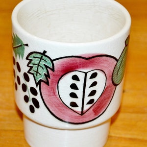 May include: White ceramic cup with a red, pink, and green fruit design. The cup has a wide rim and a base with a slightly flared lip.