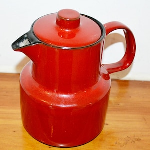 Könnte beinhalten: Eine rote Emaille-Kaffeekanne mit Deckel und Griff. Die Kanne hat einen abgerundeten Körper und einen Ausguss mit schwarzem Rand. Der Griff ist gebogen und der Deckel hat einen kleinen Knopf. Die Kanne steht auf einer Holzoberfläche.