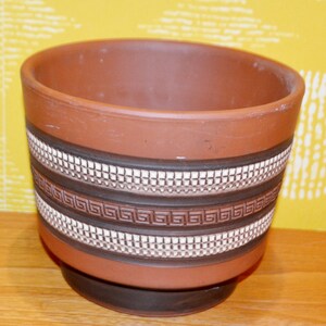 Könnte beinhalten: Ein brauner Keramik-Blumentopf mit einem dekorativen Band mit einem weißen geometrischen Muster und einem dunkelbraunen griechischen Schlüsseldesign. Der Topf hat eine breite Öffnung und einen dunkelbraunen Sockel.