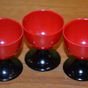 May include: Three red plastic cups with black bases. The cups have a rounded bowl shape and sit atop a short, circular black stem. The cups are arranged in a row, showcasing a retro design. The cups are likely for serving.