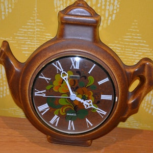 May include: A brown ceramic teapot shaped clock with Roman numeral markings and a floral design on the face. The clock face says "Quartz".