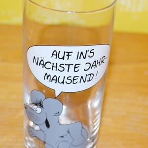 Puede incluir: Un vaso de cristal transparente con el gráfico de un ratón gris y un bocadillo blanco que contiene el texto negro AUF IN'S NÄCHSTE JAHR MAUSEND! El ratón aparece con los ojos muy abiertos y una expresión sonriente bajo el bocadillo.