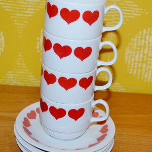 May include: A stack of three white ceramic cups with red heart patterns, each featuring a side handle, resting on a small stack of matching white saucers decorated with a border of red hearts around the rim of each individual saucer.