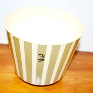 May include: A vintage wastebasket with a cream-colored interior and exterior with vertical olive green and white stripes. A small black and white logo is on the side. The wastebasket is on a wooden surface.