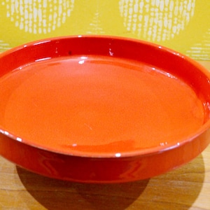 May include: A round, orange ceramic dish with a slightly raised rim. The dish is a vibrant shade of orange and appears to be glossy. The dish is sitting on a wooden surface.