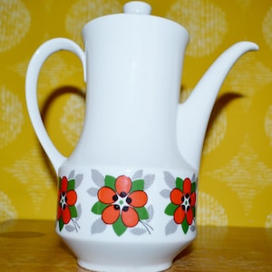 Könnte beinhalten: Weiße Kaffeekanne aus Keramik mit Blumenmuster. Das Muster zeigt rote, orange und grüne Blumen auf weißem Hintergrund.