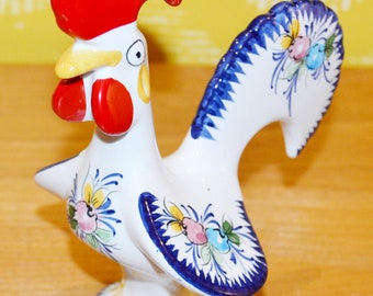 May include: A white ceramic rooster figurine with blue and yellow floral designs. The rooster has a red comb and wattle and a blue and white tail.