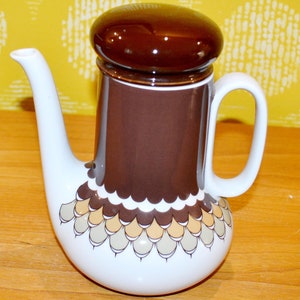 Könnte beinhalten: Eine weiße Teekanne aus Keramik mit einem braunen Deckel und einem braunen und weißen geometrischen Muster. Die Teekanne hat einen Ausguss und einen Griff.