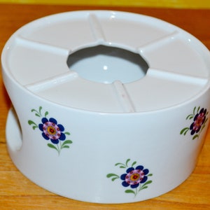 Könnte beinhalten: Weiße Keramik-Teekanne mit Blumenmuster. Die Teekanne hat einen runden Boden mit einem Loch in der Mitte für eine Teekanne. Das Blumenmuster zeigt blaue, rosa und grüne Blumen.