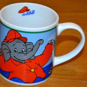 Könnte beinhalten: Eine weiße Keramiktasse mit einem Cartoon-Elefanten, der eine orangefarbene Jacke und einen Hut trägt. Der Elefant ist vor einem blauen Hintergrund mit einem grünen und blauen Rand platziert. Die Tasse hat einen weißen Henkel und den Text "förööö" im Inneren des Rands.