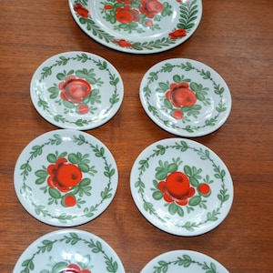 Peut inclure: Ensemble de sept assiettes en céramique blanche avec un motif floral. Les assiettes présentent des roses rouges et des feuilles vertes sur un fond blanc. Les assiettes sont disposées en cercle sur une surface en bois.