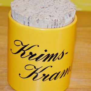 May include: A yellow ceramic jar with a cork lid. The jar has black cursive text that reads "Krims-Kram". The jar is cylindrical and sits on a wooden surface.