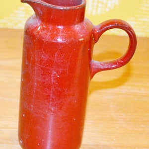 May include: A red ceramic pitcher with a handle. The pitcher has a slightly textured surface.