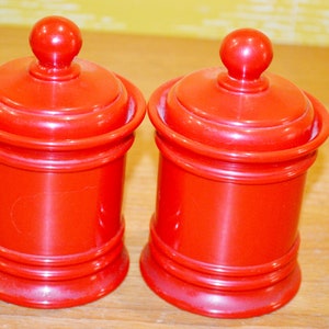 Peut inclure: Deux pots en céramique rouge avec des couvercles. Les pots ont une finition brillante et sont décorés d'une simple bordure surélevée autour du haut et du bas.