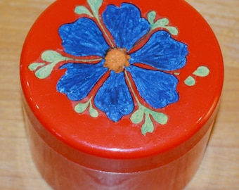 May include: A round, red container with a blue flower design on the lid. The flower has an orange center and green leaves. The container is likely made of plastic or a similar material.