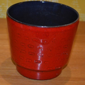 Pode incluir: Um vaso de cerâmica vermelho com um esmalte azul escuro no interior. O vaso tem uma superfície texturizada com um padrão repetitivo de pequenos quadrados.