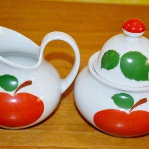 Peut inclure: Un ensemble de crémier et de sucrier en céramique blanche avec des motifs de pommes rouges et de feuilles vertes.