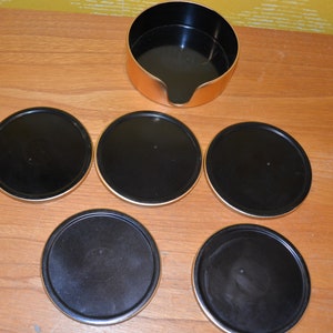 May include: Set of six black and gold coasters with a matching gold container. The coasters are round and have a black surface with a gold rim.