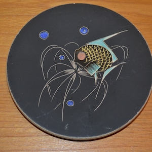 Op de afbeelding: Een zwarte keramische onderzetter met een gestileerd visontwerp. De vis is geel met blauwe en zwarte accenten en is omgeven door witte lijnen die lijken op zeewier. Er zijn blauwe stippen verspreid rond de vis.