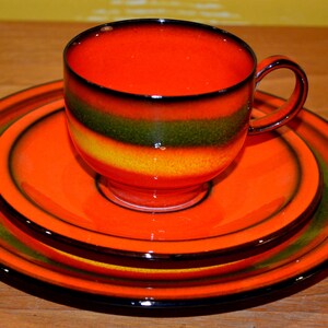 May include: A set of three orange and green ceramic plates with a cup. The plates have a concentric ring design with a black rim. The cup has a black rim and a green and yellow stripe.