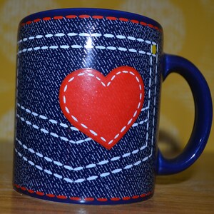 May include: A blue ceramic mug with a denim pattern and a red heart with white stitching. The mug has a white rim and a handle.