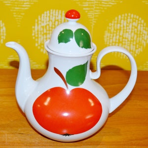 Peut inclure: Théière en céramique blanche avec un motif de pomme rouge et des feuilles vertes. La théière a un couvercle avec un bouton rouge.