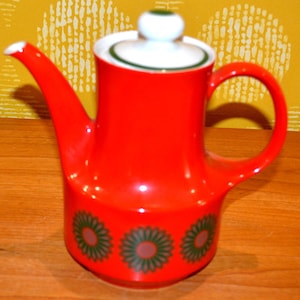 May include: Red ceramic coffee pot with a white lid and green trim. The pot has a stylized floral design in green and pink.