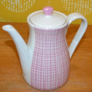 Puede incluir: Una tetera de cerámica blanca y rosa con tapa y asa. La tetera tiene un estampado de cuadros rosas en la parte delantera y un pomo rosa en la tapa. El pico y el asa son blancos. La tetera está sobre una superficie de madera.