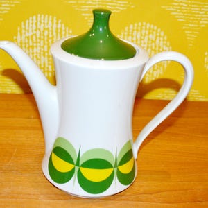 Könnte beinhalten: Weiße Keramik-Kaffeekanne mit grünem Deckel und Griff. Die Kanne hat ein dekoratives Muster aus grünen und gelben Kreisen. Der Ausguss ist lang und gebogen. Vintage-Stil.