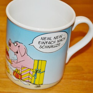 Puede incluir: Una taza de cerámica blanca con fondo azul y una ilustración de dibujos animados de dos cerdos. El texto en la taza dice "Nein, nein - einfach nach Schnauze..."