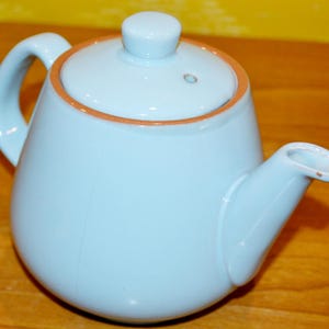Peut inclure: Une théière en céramique bleu clair avec un couvercle et un bec verseur. La théière a une bordure brune autour du haut.