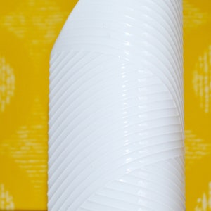 Puede incluir: Un jarrón blanco curvado con un diseño texturizado y acanalado. El jarrón tiene un acabado liso y brillante y se apoya sobre una superficie de madera sobre un fondo amarillo. La forma del jarrón recuerda a una escultura moderna.