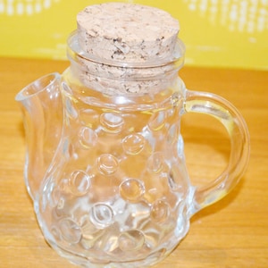 Peut inclure: Une crémière en verre transparent avec un bouchon en liège. La crémière a une poignée et un bec verseur. Le corps de la crémière est orné d'un motif de cercles en relief. Le bouchon en liège est brun clair.