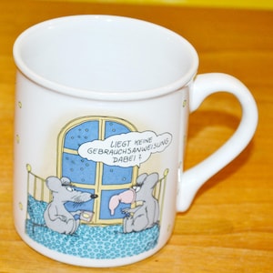 Puede incluir: Taza de cerámica blanca con una ilustración de dibujos animados de dos ratones en la cama. El texto en la taza dice "Liegt keine Gebrauchsanweisung dabei?"