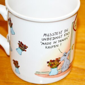 Puede incluir: Taza de cerámica blanca con asa, con una ilustración de dibujos animados de un ratón y flores. La taza tiene texto en alemán: "Musstest du unbedingt eins 'Made in Taiwan' kaufen?"